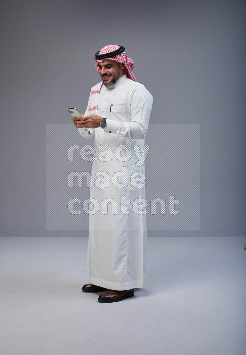 Saudi man Wearing Thob and red Shomag standing texting on phone on Gray background