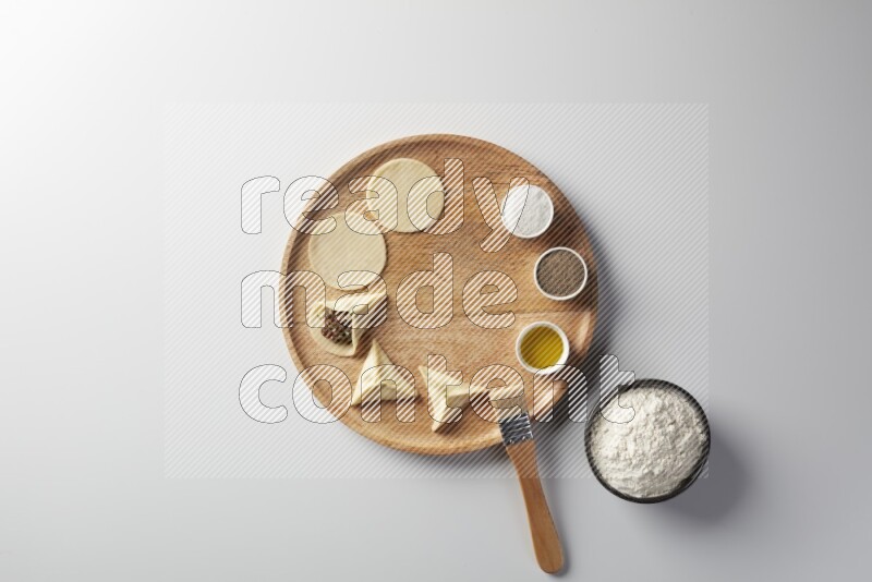 two closed sambosas and one open sambosa filled with meat while flour, salt, black pepper and oil with oil brush aside in a wooden dish on a white background