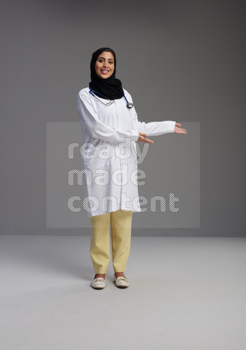 Saudi woman wearing lab coat with stethoscope standing interacting with the camera on Gray background