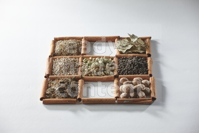 9 squares of cinnamon sticks full of cardamom in the middle surrounded by nutmeg, salt, dried basil, white pepper, cumin, allspice, bay laurel leaves and black pepper on white flooring