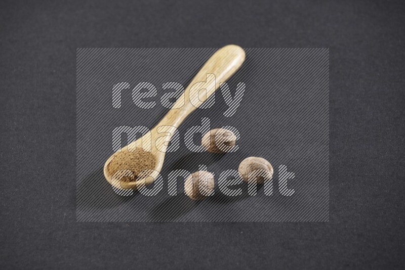 A wooden spoon full of nutmeg powder with whole nutmeg seeds beside it on a black flooring