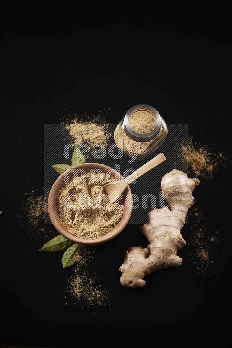 A wooden bowl full of ground ginger powder with a glass jar beside it and fresh ginger on black background