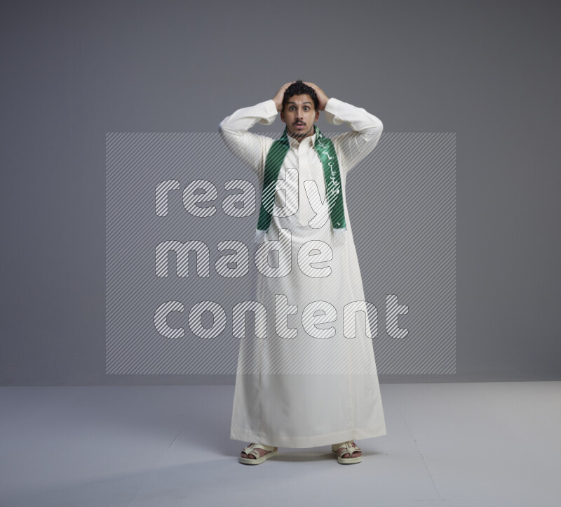 A Saudi man standing wearing thob and Saudi flag scarf on gray background