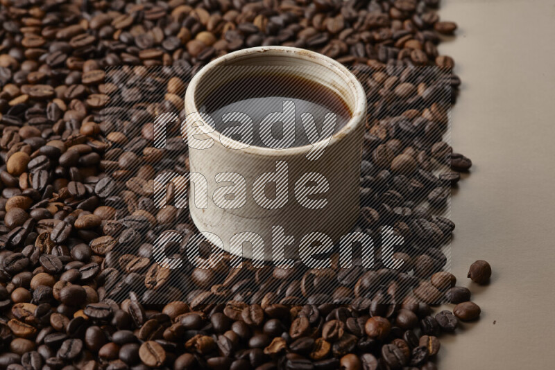 A beige pottery cup of coffee surrounded by roasted coffee beans on beige background