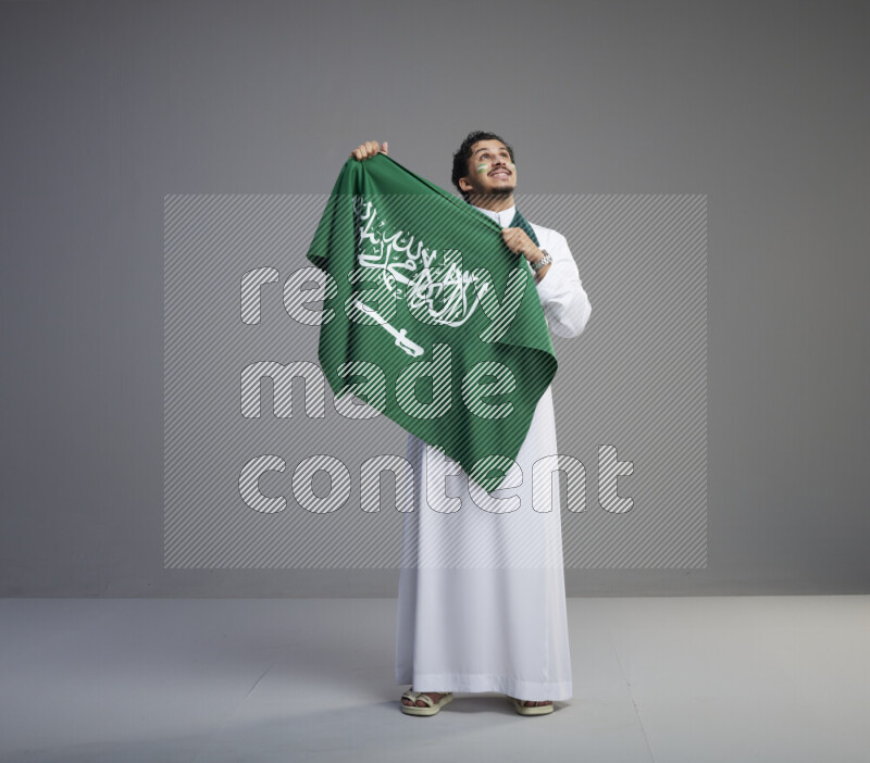 A Saudi man standing wearing thob with face painting wrapping Saudi flag scarf and holding big Saudi flag on gray background