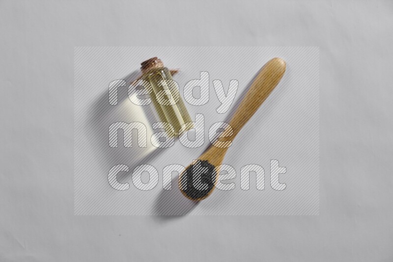 A wooden spoon full of black seeds with a glass bottle of black seeds oil on a white flooring