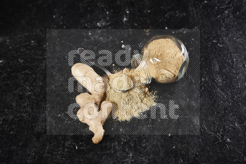 A glass jar full of ground ginger powder flipped with some spilling powder on black background