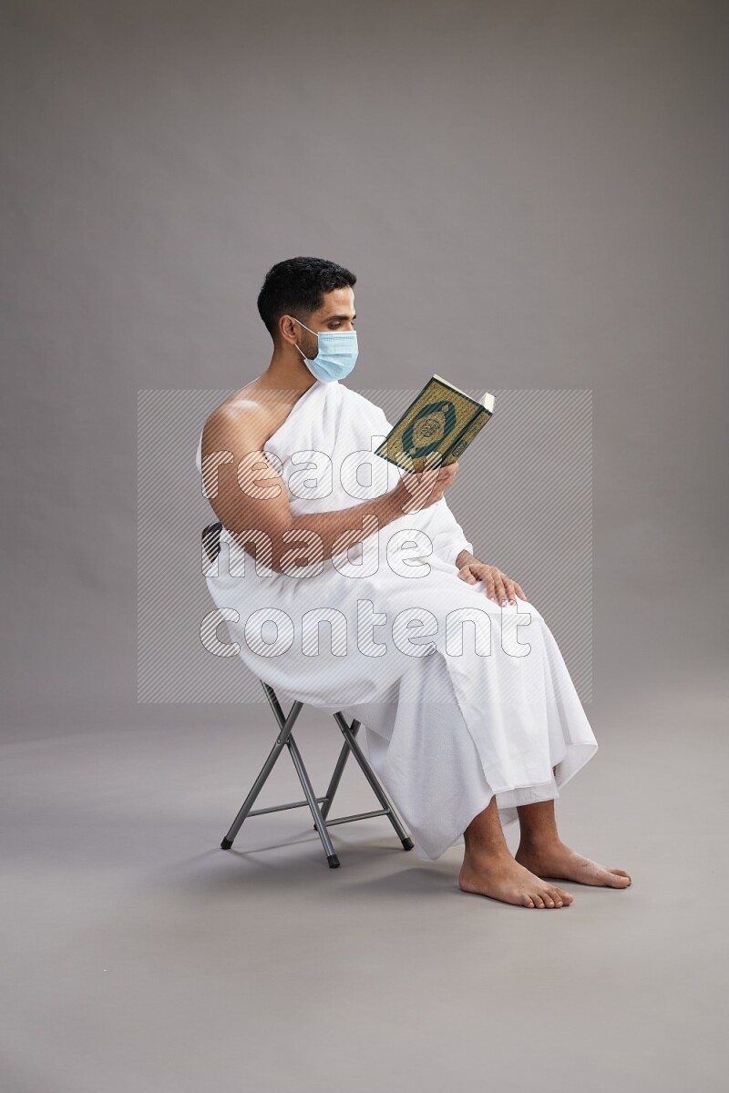 A man wearing Ehram with face mask sitting on chair reading quran on gray background