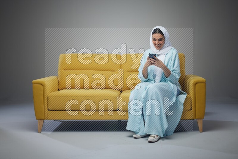 A Saudi woman wearing a light blue Abaya and white head scarf sitting on a yellow sofa and using her phone eye level on a grey background