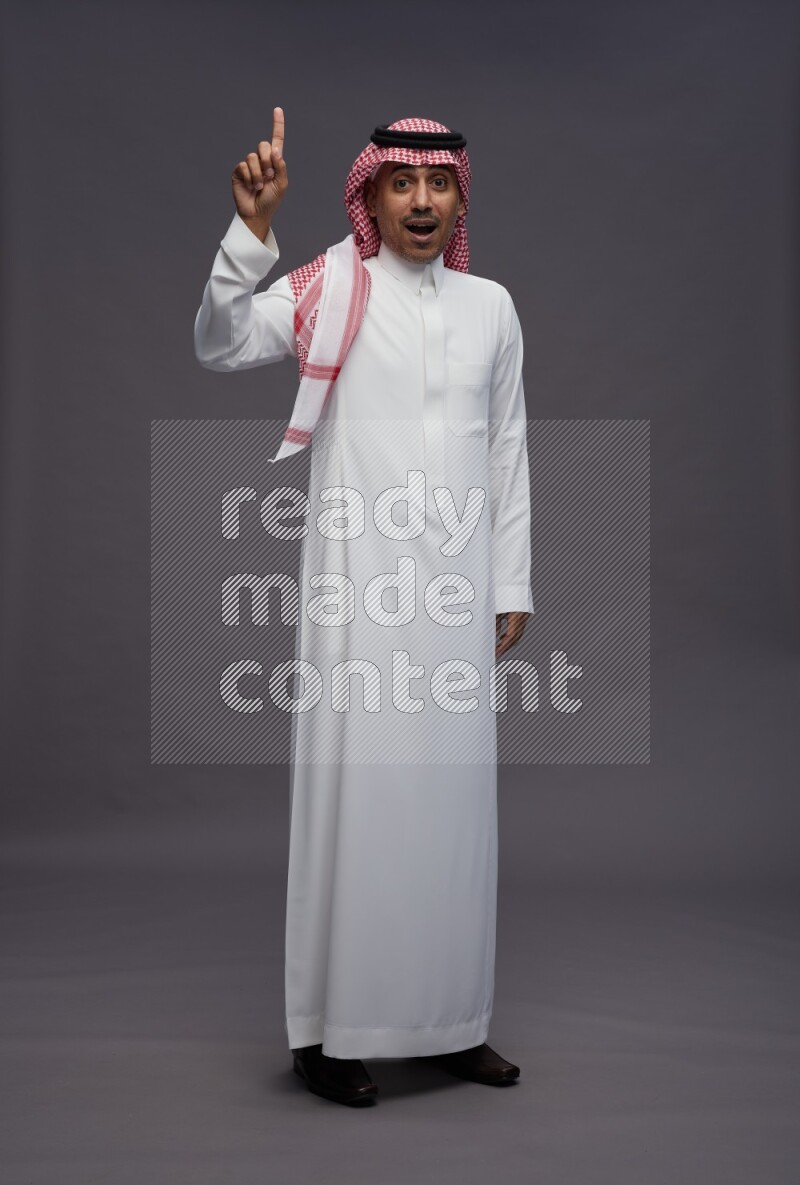 Saudi man wearing thob and shomag standing interacting with the camera on gray background