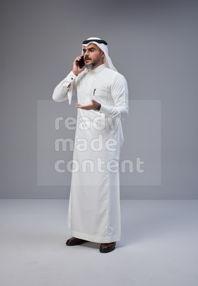 Saudi man Wearing Thob and white Shomag standing talking on phone on Gray background
