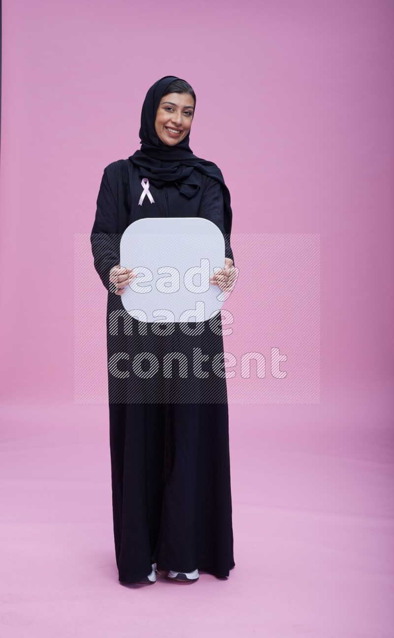 Saudi woman wearing pink ribbon on Abaya standing holding social media signs on pink background