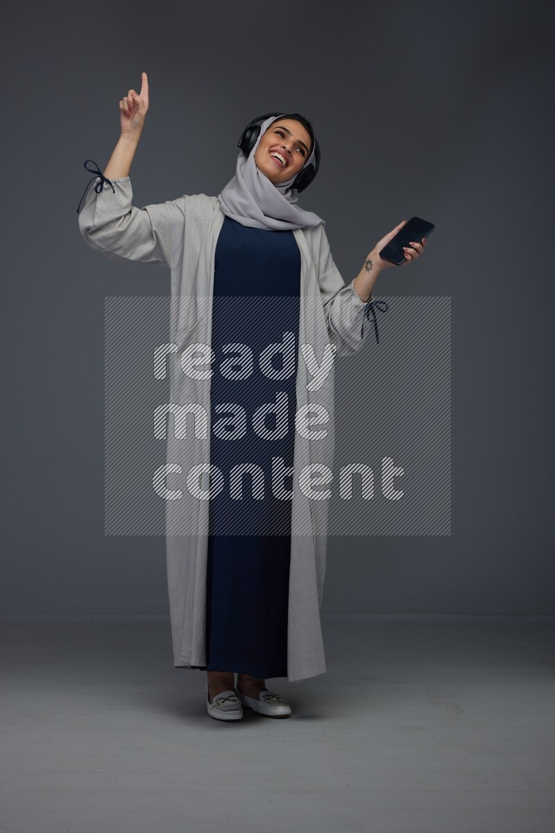 A Saudi woman wearing a light gray Abaya and head scarf standing and listening to music on a grey background