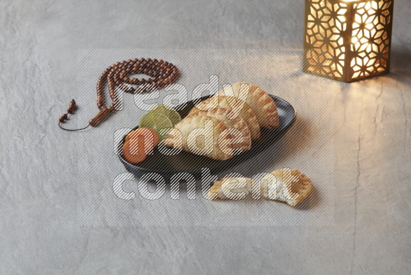 Four fried sambosas in an oval shaped black plate, beside a cut cheese sambosa, a brown misbaha and a golden lantern on a gray background