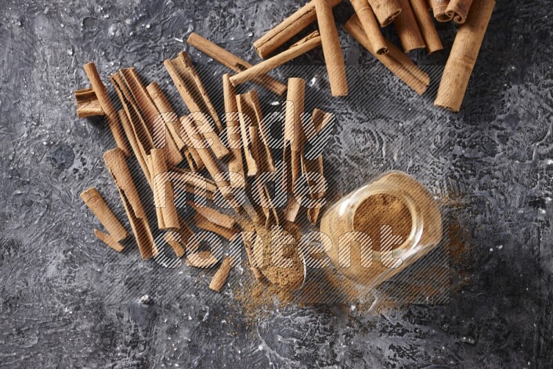 Herbal glass jar and a metal spoon full of cinnamon powder surrounded by cinnamon sticks on textured black background
