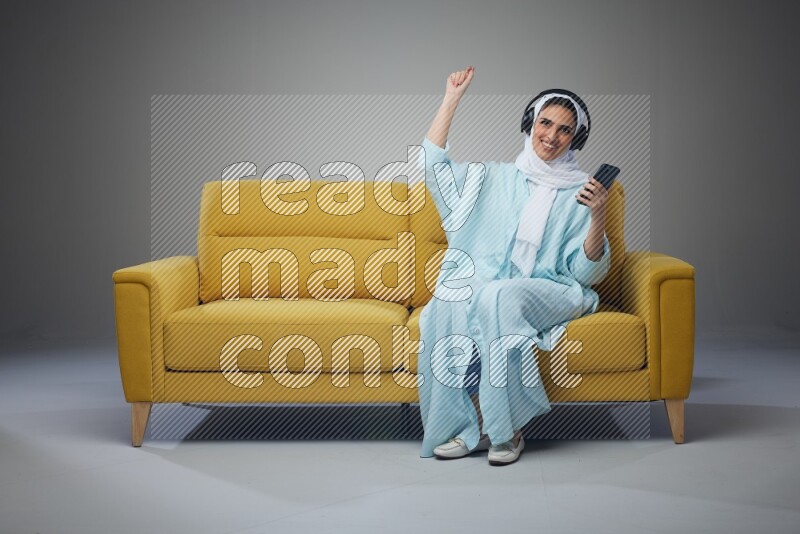 A Saudi woman wearing a light blue Abaya and a white head scarf sitting on a yellow sofa and holding an iPad while wearing headphone eye level on a grey background