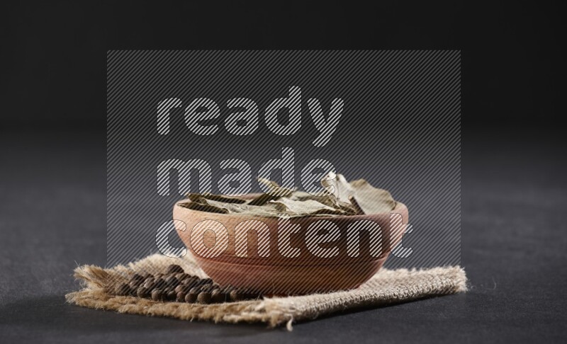 A wooden bowl filled with dried bay leaves on a piece of burlap with bunch of allspice berries on black flooring