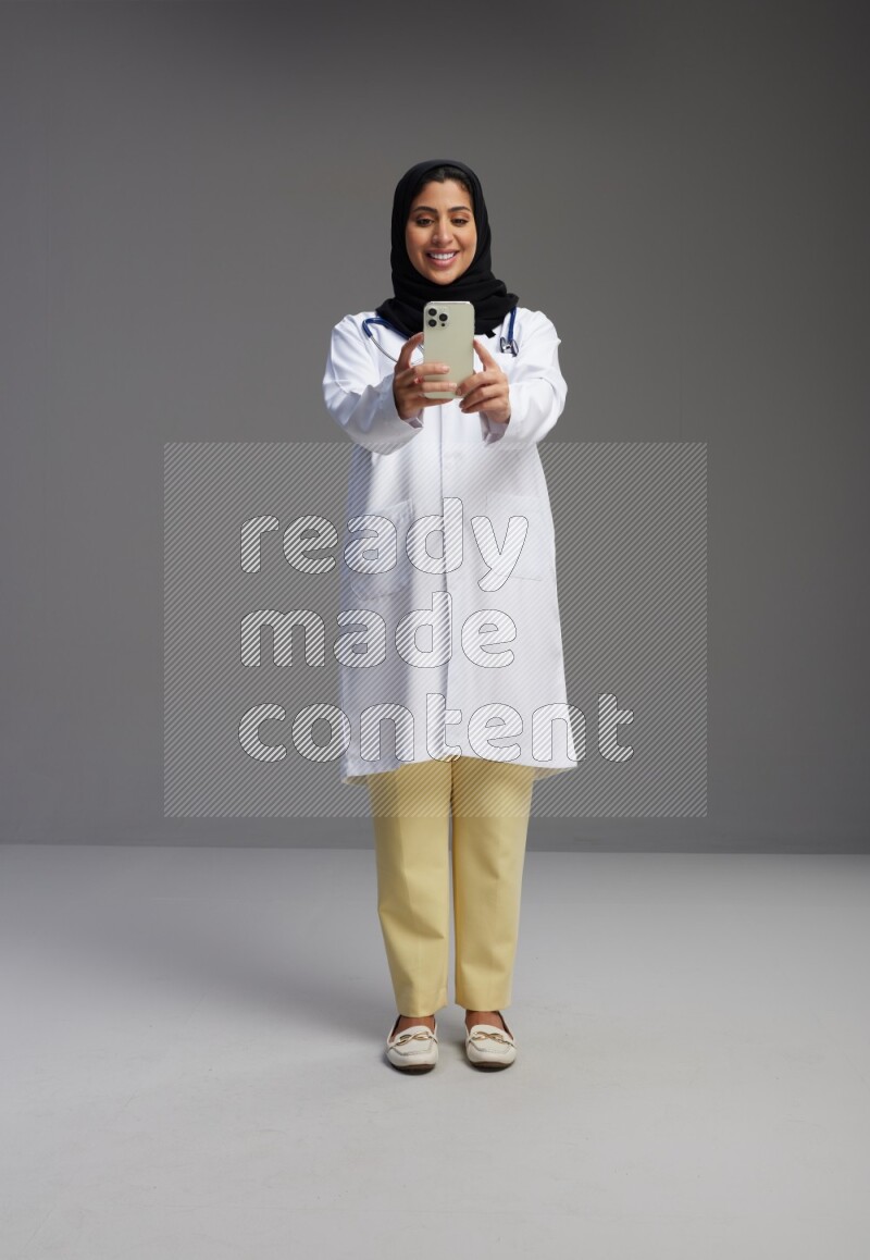 Saudi woman wearing lab coat with stethoscope standing taking selfie on Gray background
