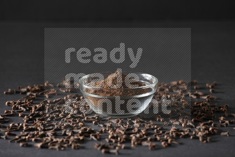 وعاء زجاجي ممتلئ ببودرة القرنفل مع حبوب القرنفل المتناثرة على أرضية سوداء