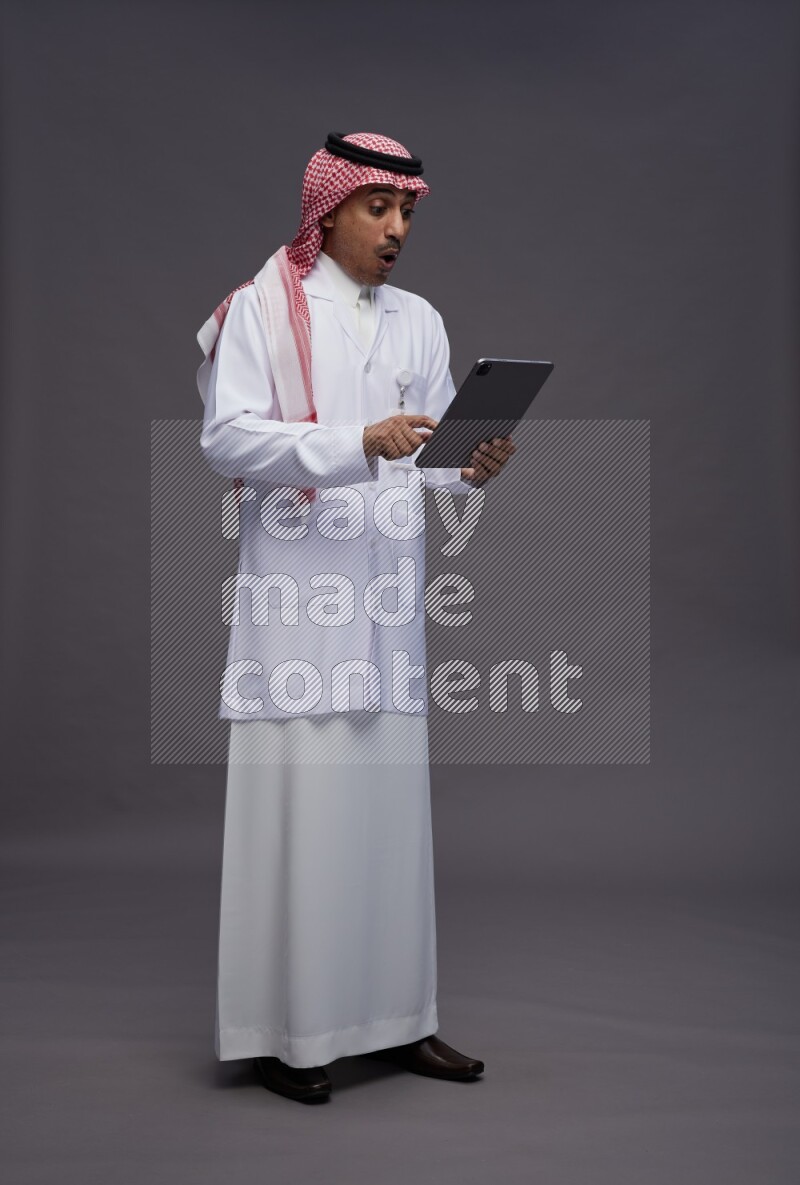 Saudi man wearing thob with lab coat and shomag with pocket employee badge standing working on tablet on gray background