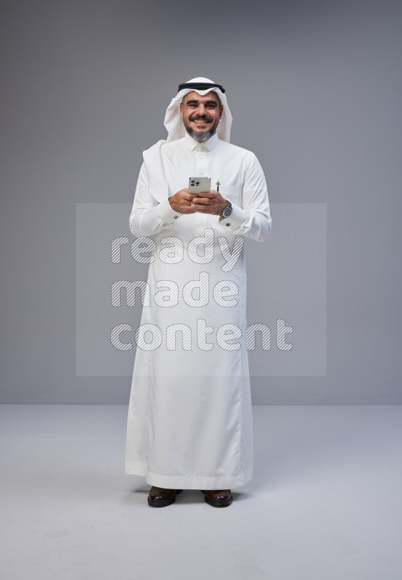 Saudi man Wearing Thob and white Shomag standing texting on phone on Gray background