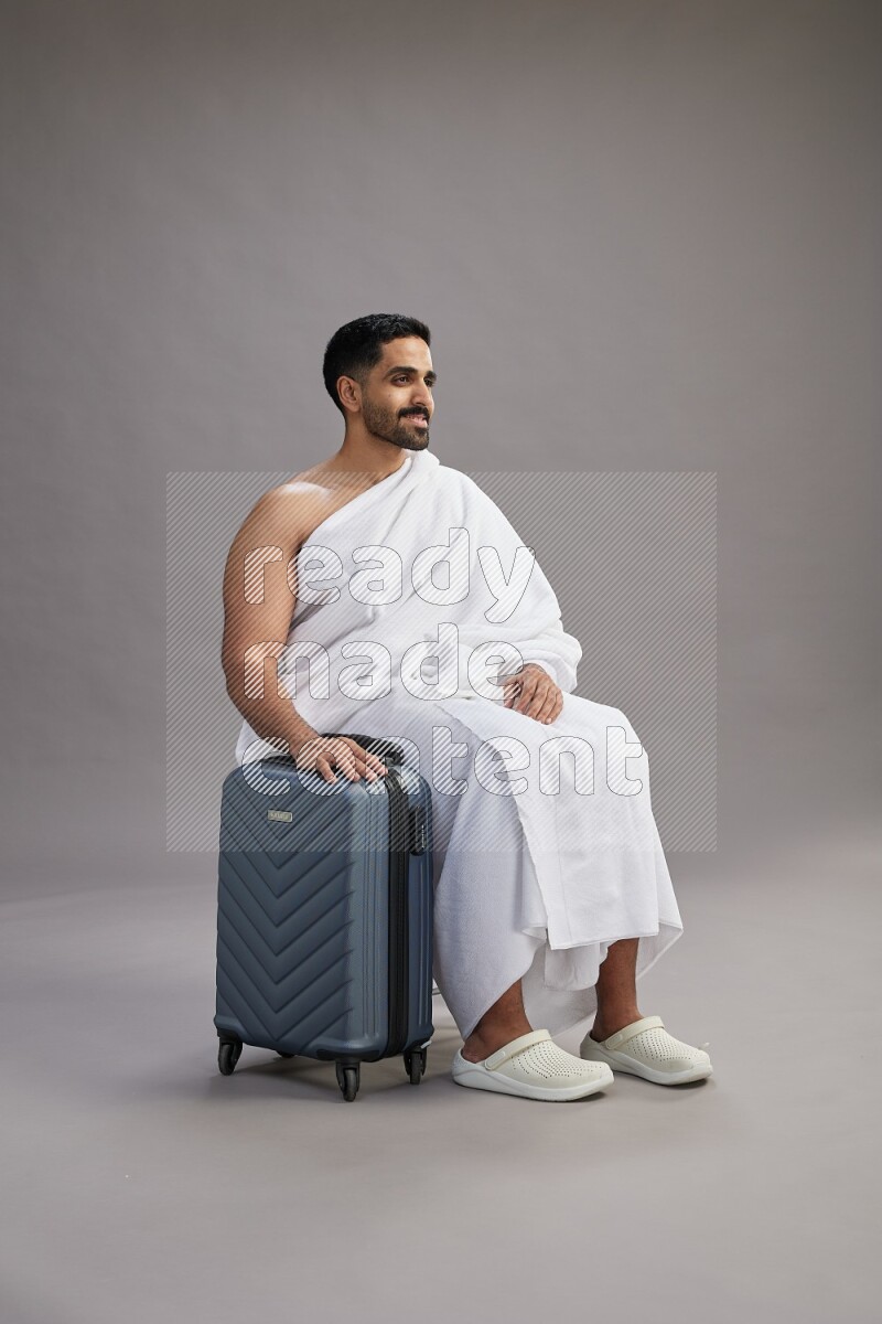 A man wearing Ehram sitting on chair holding traveling bag on gray background