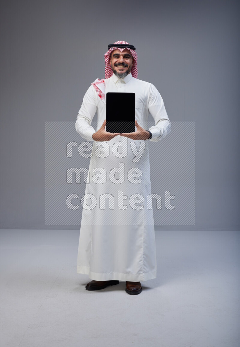 Saudi man Wearing Thob and red Shomag standing showing tablet to camera on Gray background