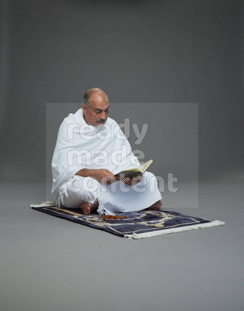 A man wearing Ehram sitting on mate prayer reading quran on gray background