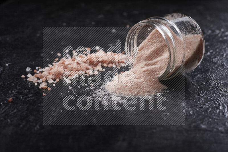 A glass jar full of fine himalayan salt with some himalayan crystals beside it on a black background