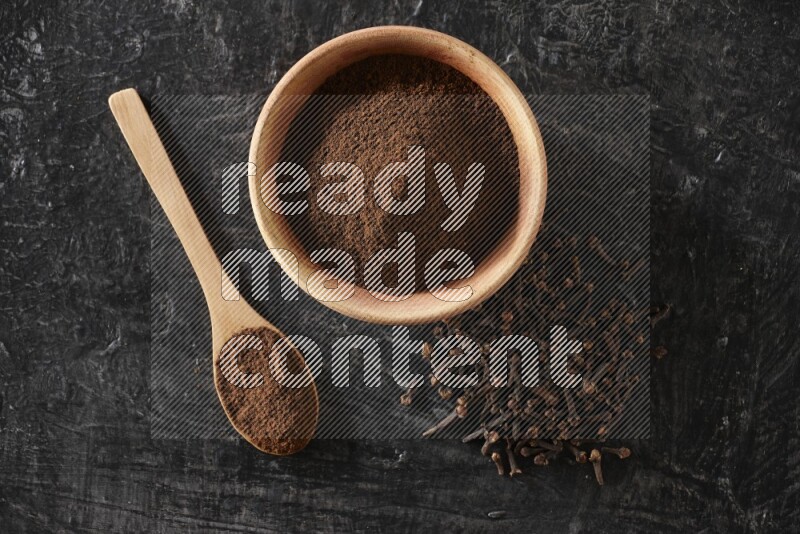 A wooden bowl and a wooden spoon full of cloves powder with spreaded cloves on a textured black flooring