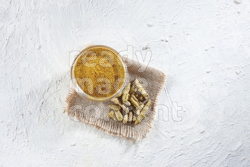 A glass bowl full of turmeric powder and dried turmeric whole finger on a piece of burlap on a textured white flooring