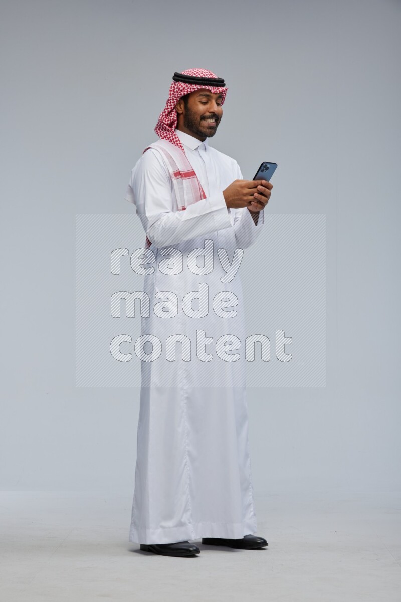 Saudi man Wearing Thob and shomag standing texting on phone on Gray background