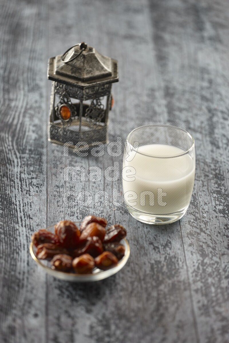 A silver lantern with different drinks, dates, nuts, prayer beads and quran on grey wooden background