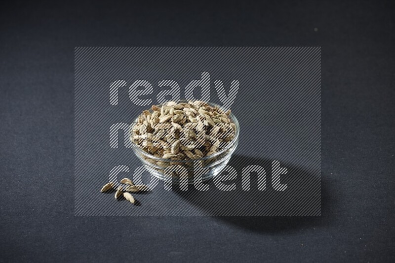 A glass bowl full of cardamom and more seeds spreaded beneath the bowl on black flooring
