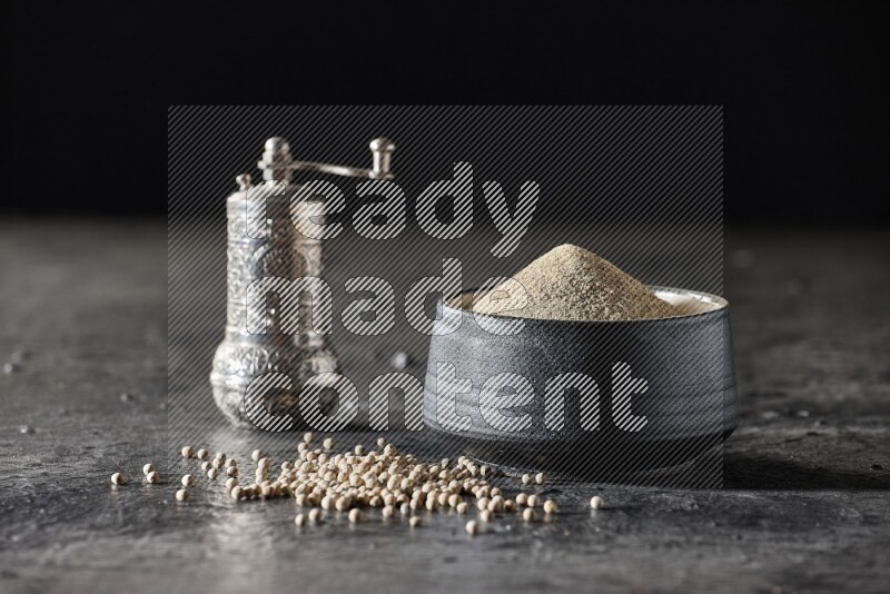 Black pottery bowl full of white pepper powder with pepper beads and metal pepper grinder on textured black flooring