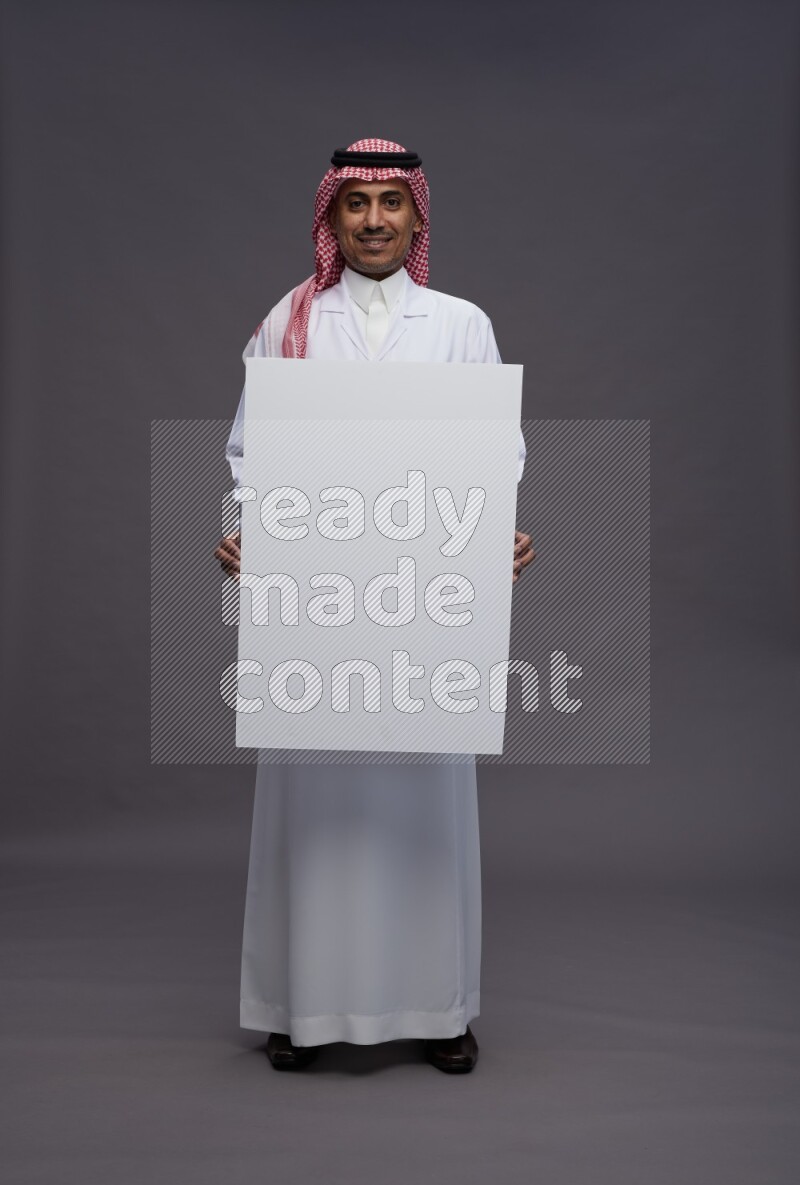 Saudi man wearing thob with lab coat and shomag with pocket employee badge standing holding board on gray background