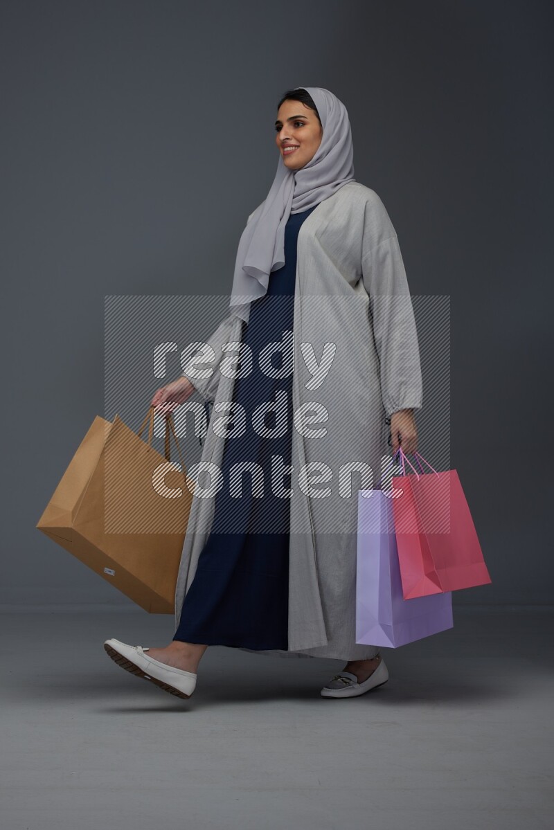 A Saudi woman wearing a light gray Abaya and head scarf standing and holding shopping bags on a grey background