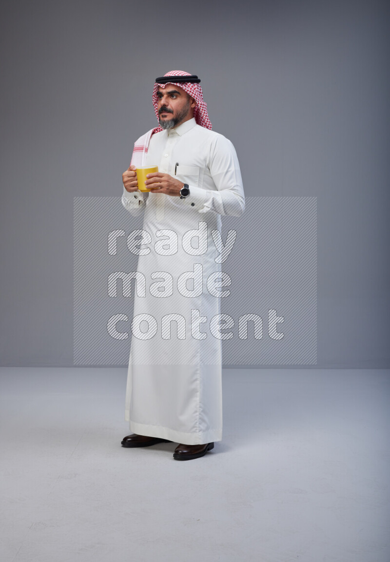 Saudi man Wearing Thob and red Shomag standing holding mug on Gray background