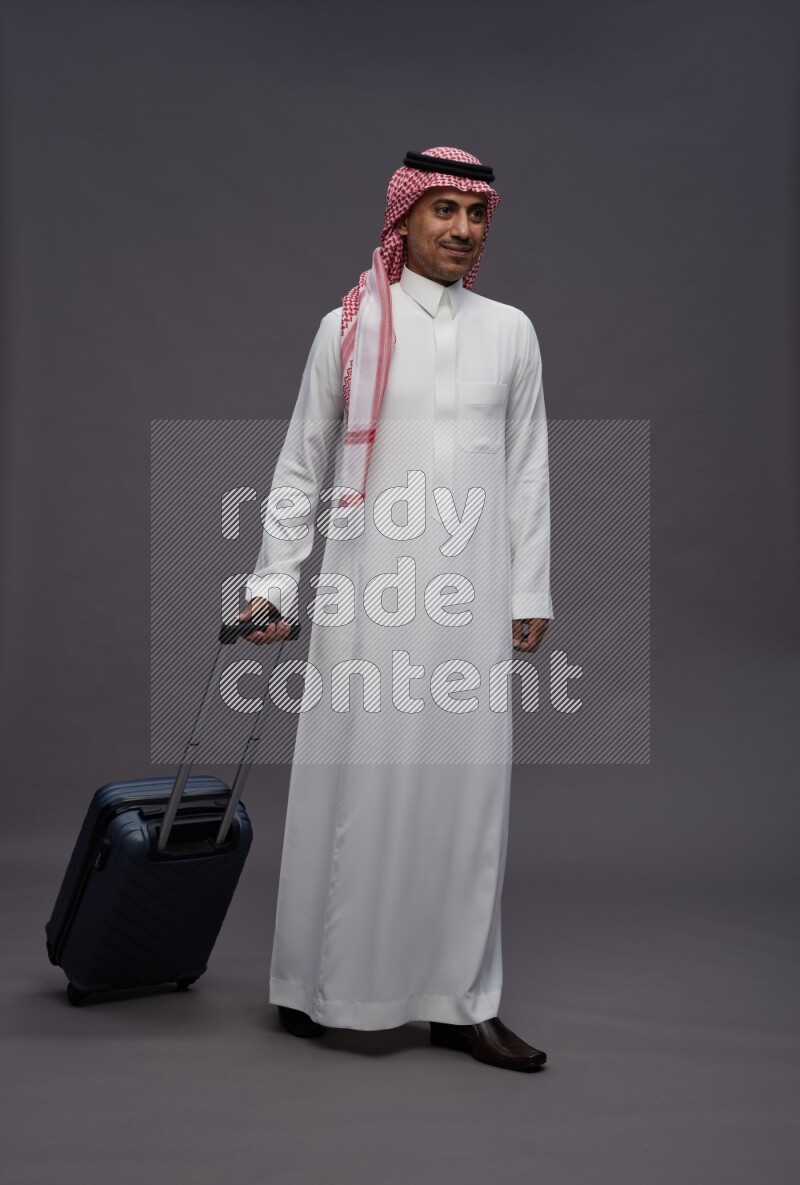 Saudi man wearing thob and shomag standing holding bag on gray background
