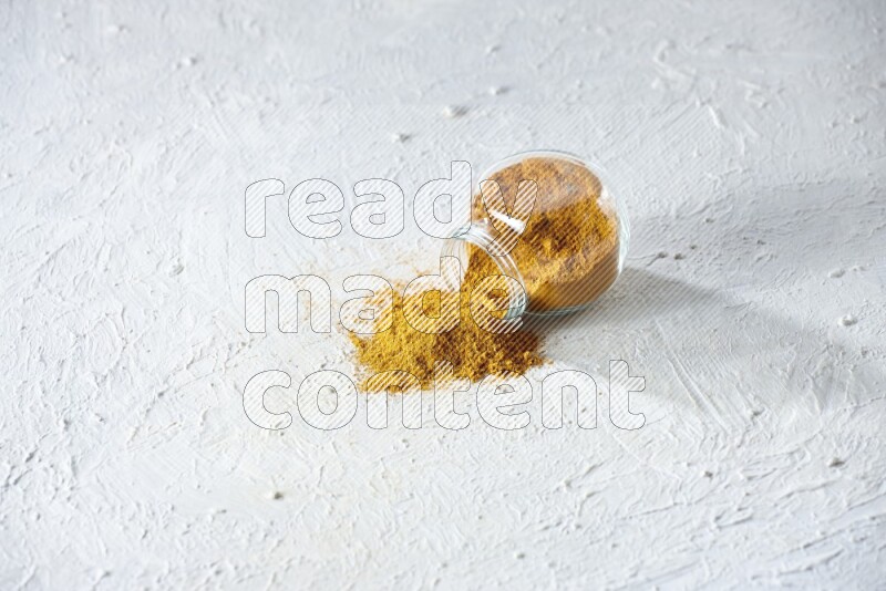 A flipped glass spice jar full of turmeric powder and powder fell out it on textured white flooring