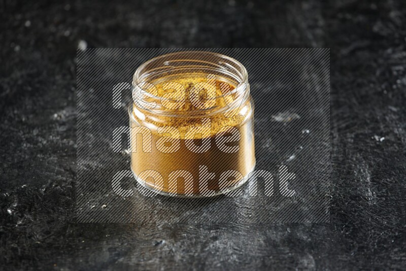 A glass jar full of turmeric powder on a textured black flooring