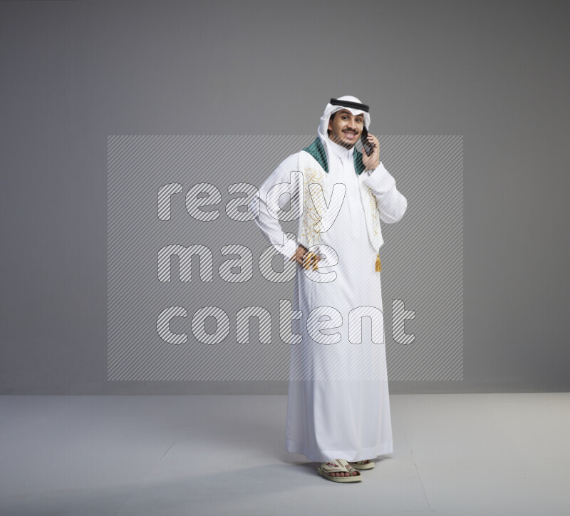 A saudi man standing wearing thob and white shomag with flag scarf on his neck talking on phone on gray background