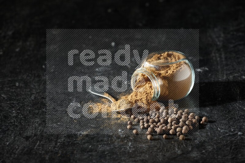 وعاء زجاجي للتوابل مقلوب وملعقة معدنية ممتلئان ببودرة بهار الفلفل الحلو والبودرة تناثرت من الوعاء مع بعض الحبوب على خلفية سوداء