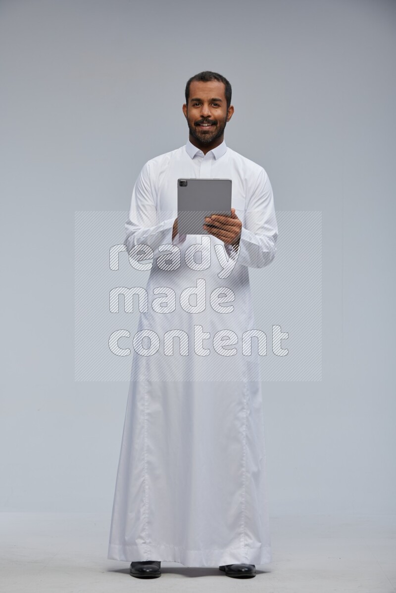 Saudi man wearing Thob standing working on tablet on Gray background