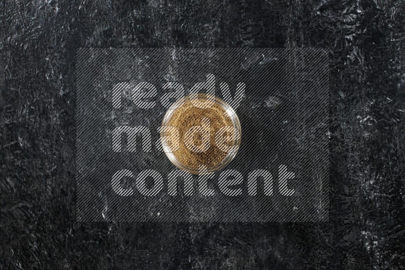A glass jar full of cumin powder on a textured black flooring