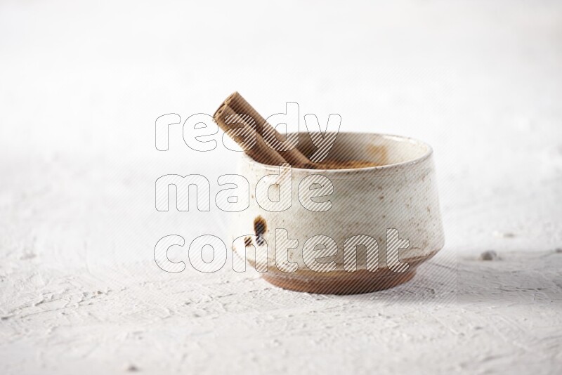 Ceramic beige bowl full of cinnamon powder with a cinnamon stick on a textured white background