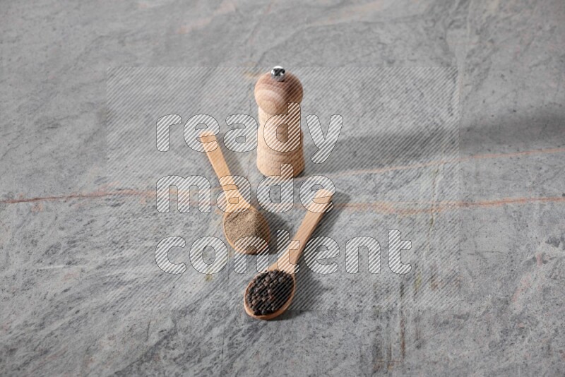 2 wooden spoons, one full of black pepper powder and the other full of black pepper beads and wooden grinder on a marble flooring