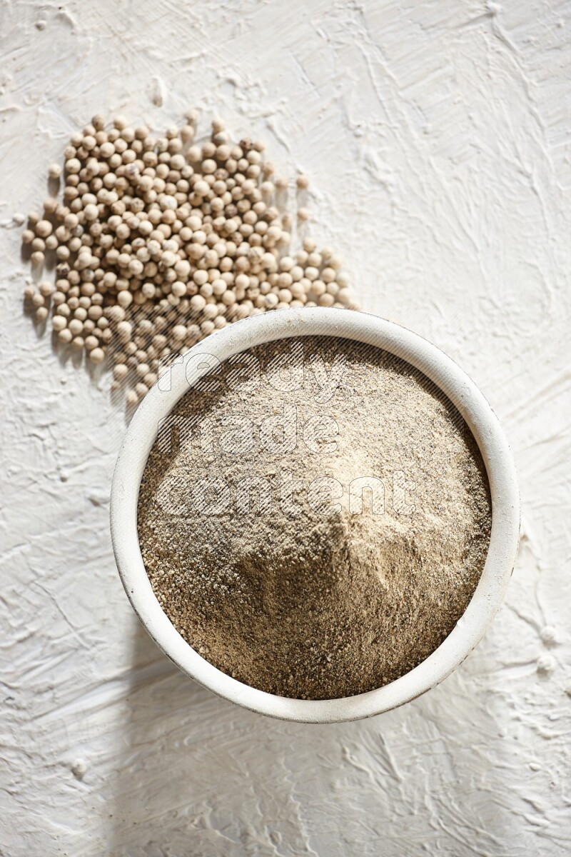 white pottery bowl full of white pepper powder with pepper beads on textured white flooring
