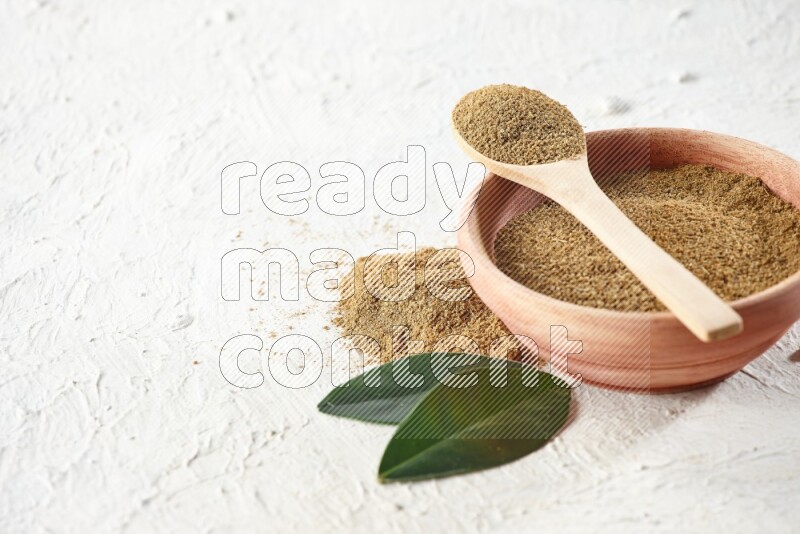 A wooden bowl and wooden spoon full of cumin powder on textured white flooring