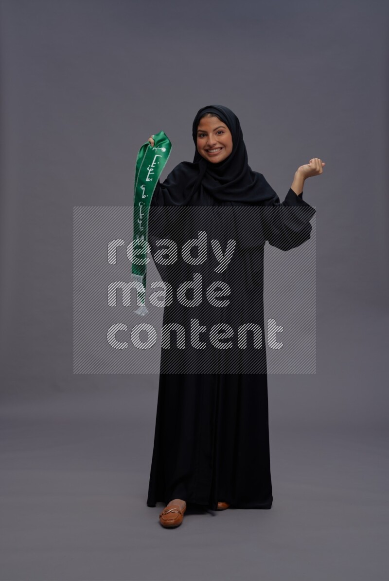 Saudi woman wearing Abaya standing holding Saudi flag on gray background
